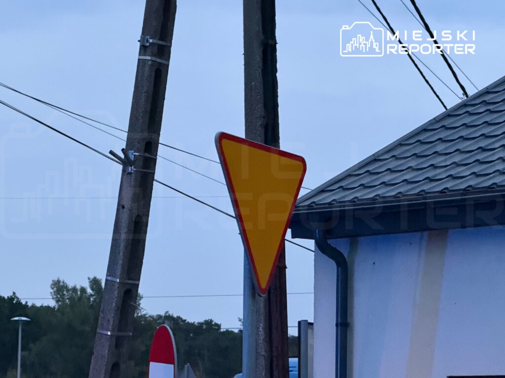 Żółty znak ostrzegawczy umieszczony na słupie, w tle widoczny budynek z dachówką.