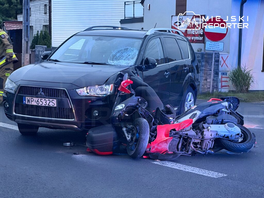 Czarny samochód osobowy z uszkodzoną karoserią stoi obok przewróconego motocykla na drodze.