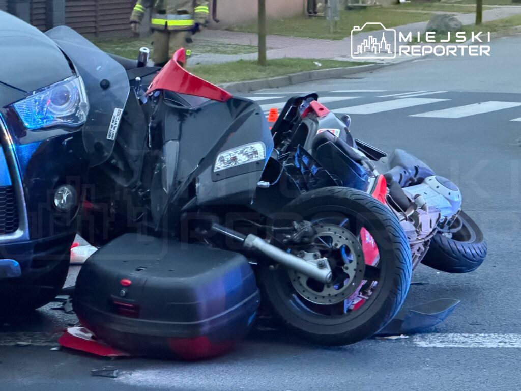 Motocykl leżący na boku, zderzony z czarnym samochodem osobowym na drodze. W tle widać strażaków.