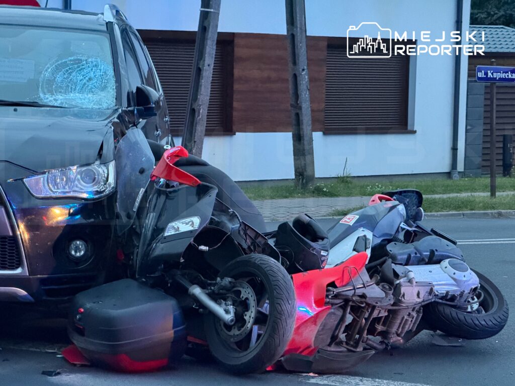 Zniszczony motocykl leży na jezdni obok uszkodzonego czarnego SUV-a, w tle widoczny budynek i znak drogowy.