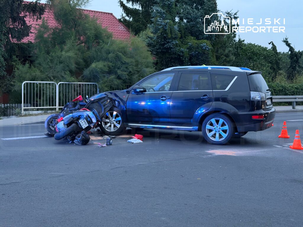 Motocykl leży przewrócony obok czarnego SUV-a na drodze, otoczony przez trójkąty ostrzegawcze.