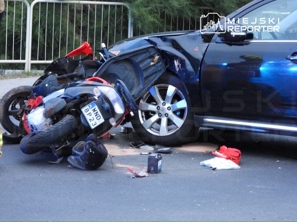 Motocykl leży na boku obok uszkodzonego samochodu osobowego, na jezdni w miejskim otoczeniu.