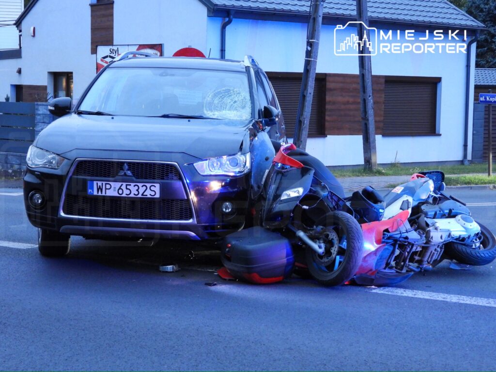 Czarny samochód osobowy z uszkodzoną szybą stoi na drodze obok przewróconego motocykla.