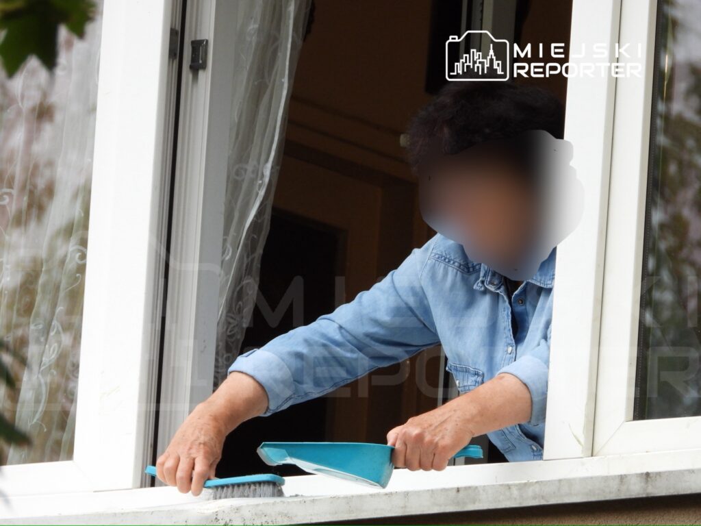 Kobieta w niebieskiej koszuli sprząta parapet okna, używając szczotki i szufelki.