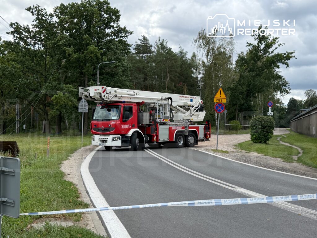 Czerwony wóz strażacki z podnośnikiem umiejscowiony na zakręcie drogi, otoczony taśmą zabezpieczającą.