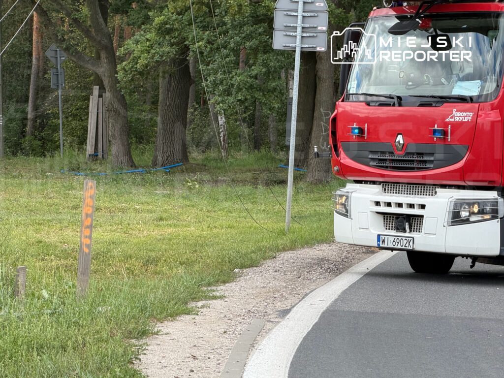 Czerwony wóz strażacki zatrzymany przy drodze, obok znaków drogowych i drzew w tle.