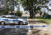 Policyjny radiowóz z niebieskimi oznaczeniami blokuje drogę, a w tle widać drzewa i budynek z szarym murem.