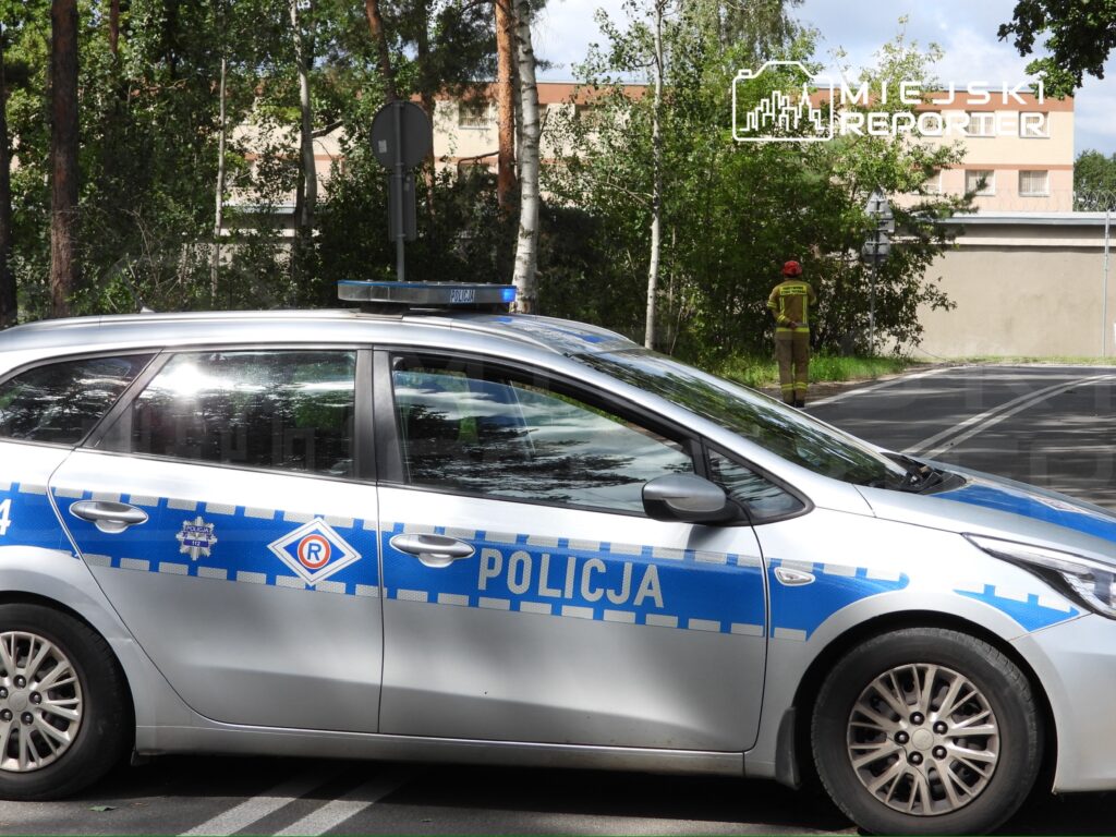 Policyjny radiowóz zaparkowany na drodze obok rosnących drzew, w tle strażak obserwujący otoczenie.