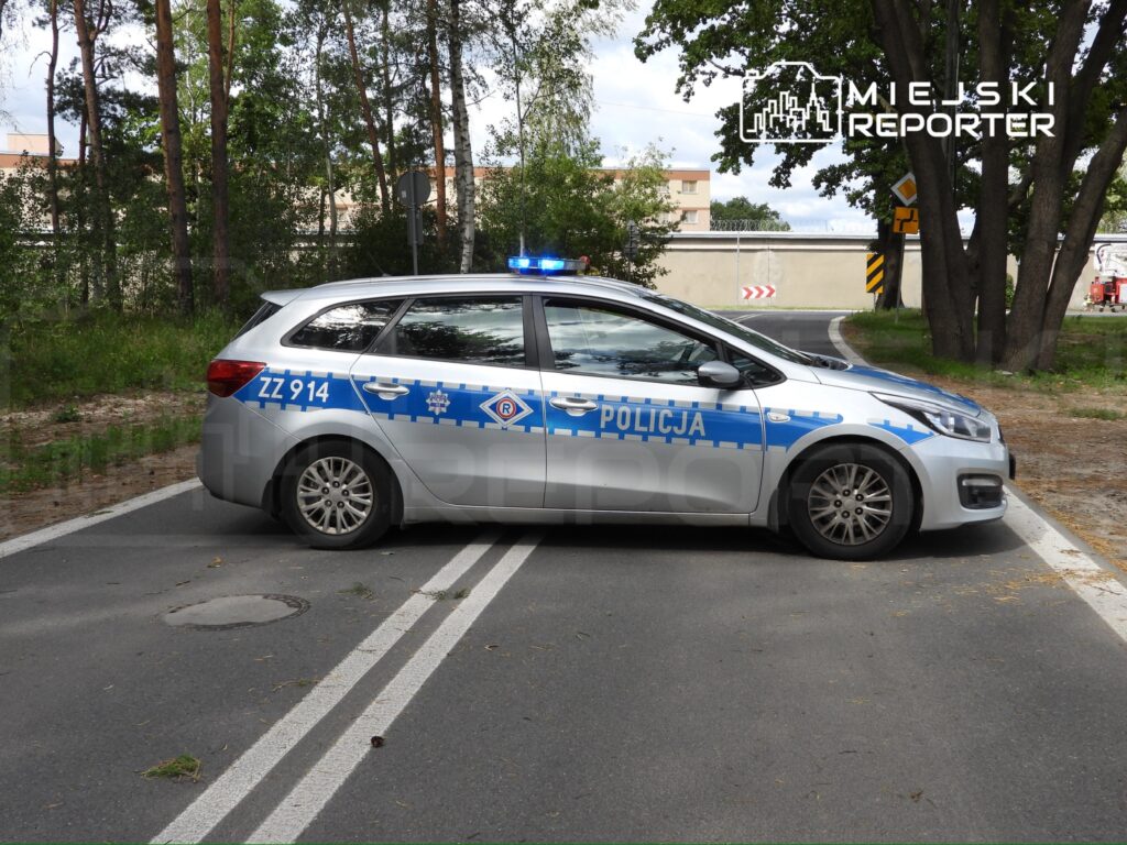 Policyjny samochód osobowy z niebieskimi światłami zatrzymany na drodze w otoczeniu drzew.