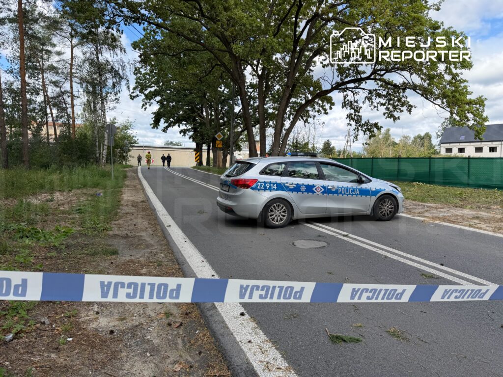 Policyjny radiowóz z zablokowanym dostępem stoi na drodze, a funkcjonariusze prowadzą czynności na poboczu w otoczeniu drzew.
