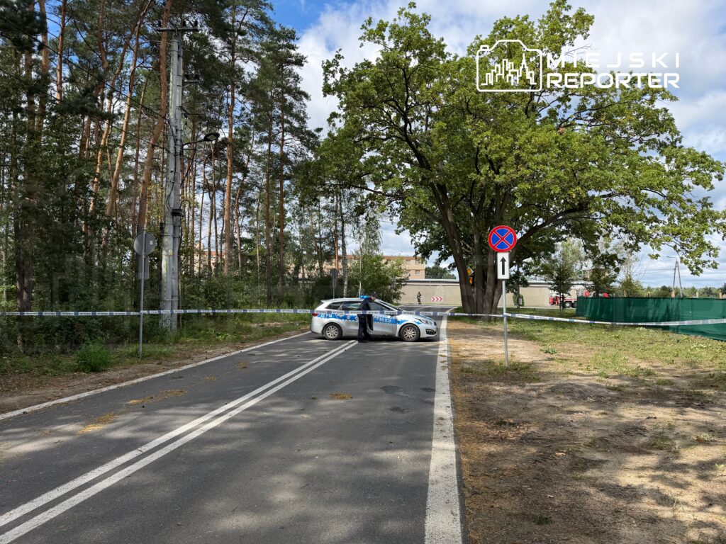 Policyjny radiowóz zabezpieczający teren przy drodze otoczonej drzewami, z oznaczeniami zakazu parkowania.