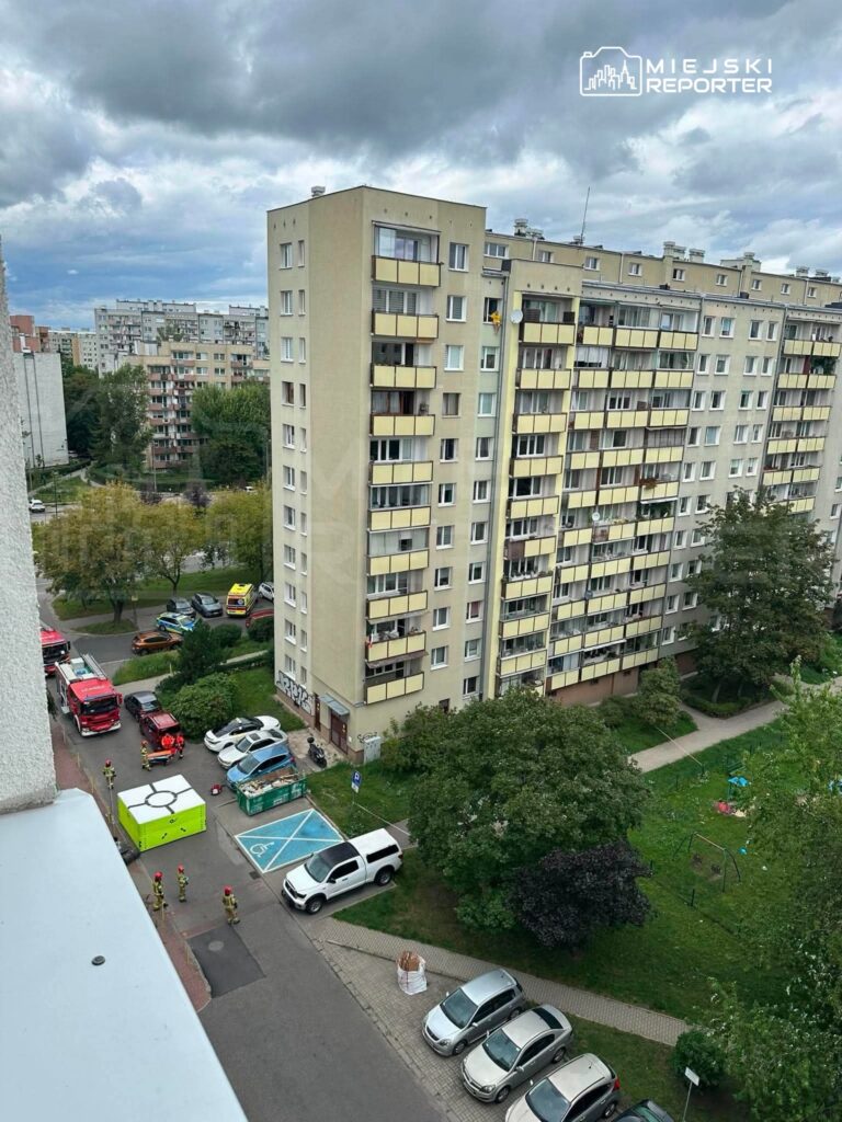Grupa strażaków w mundurach interweniuje przy zielonym pojeździe w pobliżu bloku mieszkalnego. W tle widać chmury.