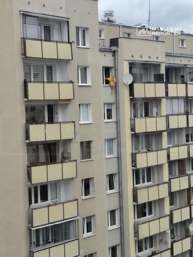 Mężczyzna w żółtej koszulce siedzący na parapecie okna w bloku mieszkalnym.