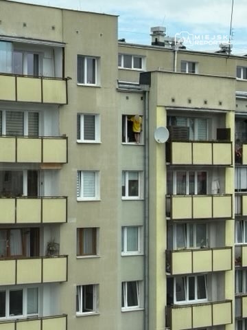 Kobieta w żółtej odzieży wychyla się przez okno w bloku mieszkalnym.