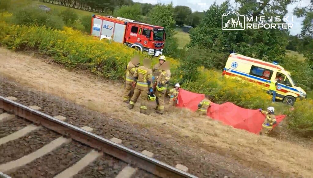 Grupa strażaków w mundurach zabezpiecza miejsce zdarzenia obok torów kolejowych, w tle widać ambulans.