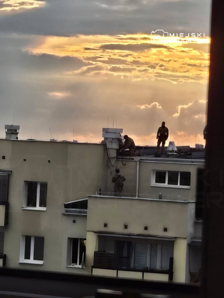 Grupa pracowników na dachu budynku wykonuje prace montażowe w tle zachodzącego słońca.