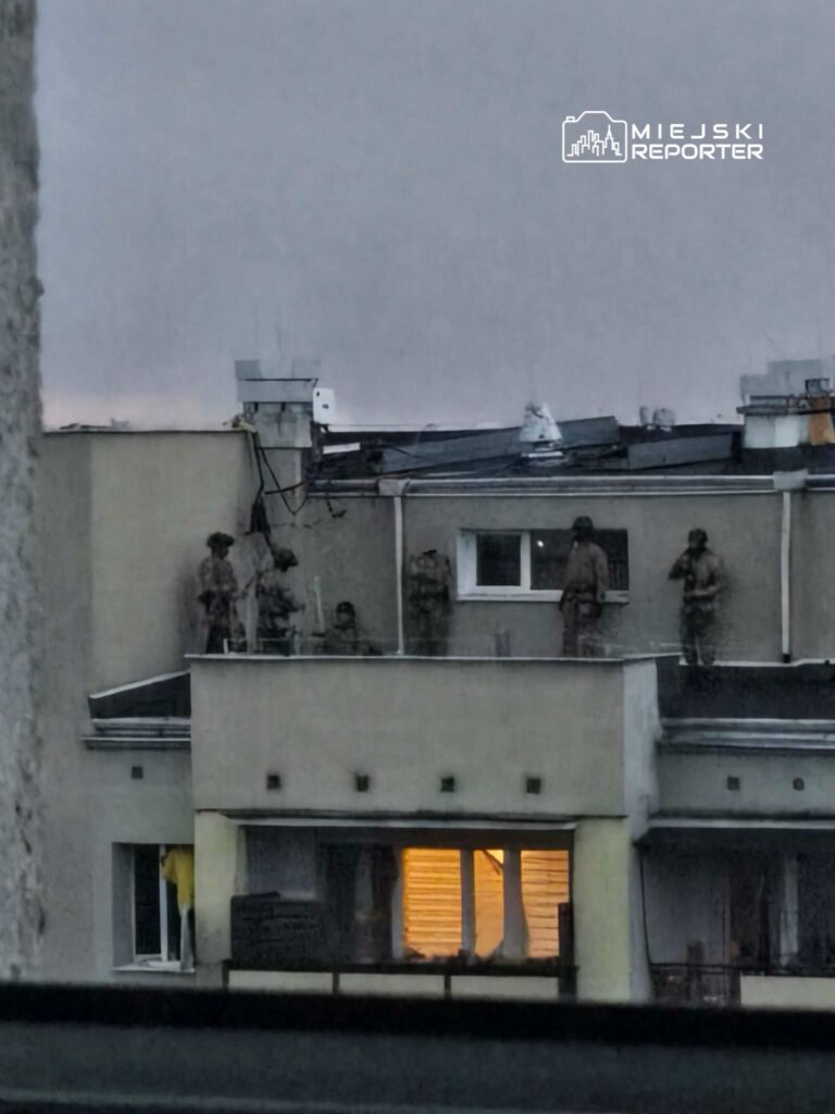 Grupa żołnierzy w mundurach stoi na dachu budynku, obserwując okolicę w szarym świetle zmierzchu.