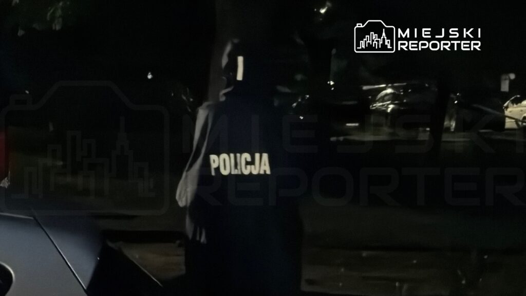 Funkcjonariusz policji w mundurze, stojący w ciemnym otoczeniu, z widocznym napisem "POLICJA" na plecach.