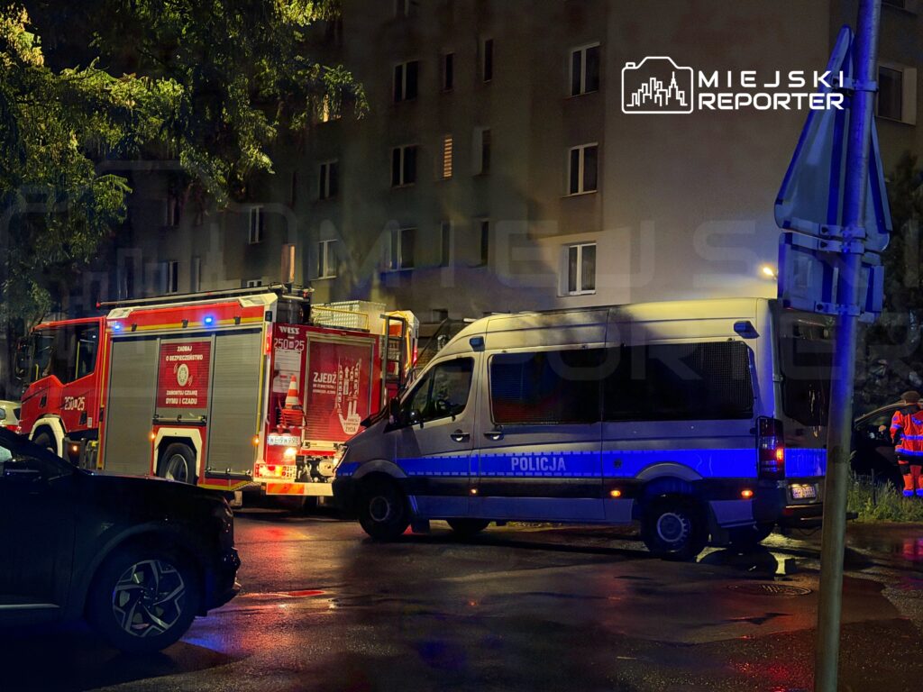 W nocy, w oświetlonym miejscu, straż pożarna i policyjny van interweniują przy budynku mieszkalnym.