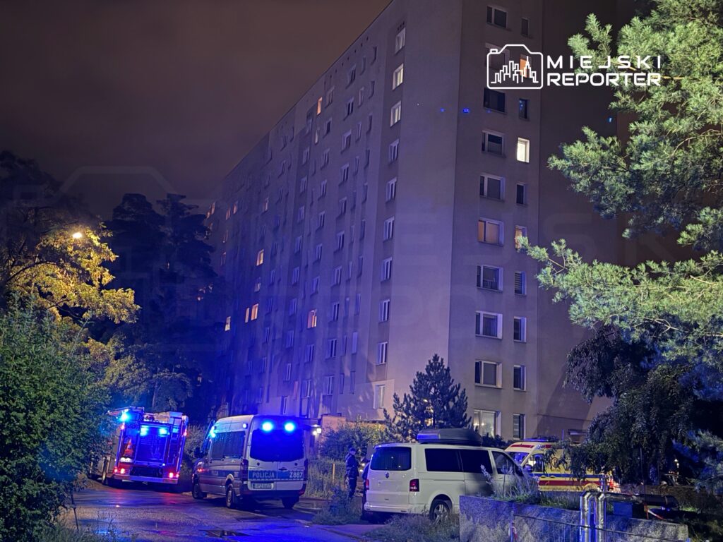 W nocy przy bloku mieszkalnym interweniują służby ratunkowe, w tym straż pożarna i policja, z włączonymi sygnalizatorami.