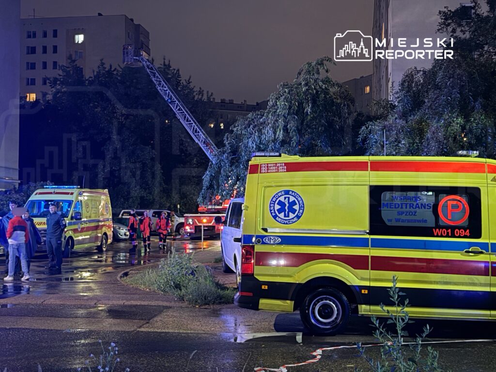 Grupa ratowników medycznych w czerwonych strojach przemieszcza się obok ambulansów na deszczowej ulicy, w tle widać drabinę strażacką.