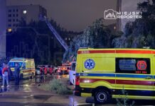 Grupa ratowników medycznych w czerwonych strojach przemieszcza się obok ambulansów na deszczowej ulicy, w tle widać drabinę strażacką.