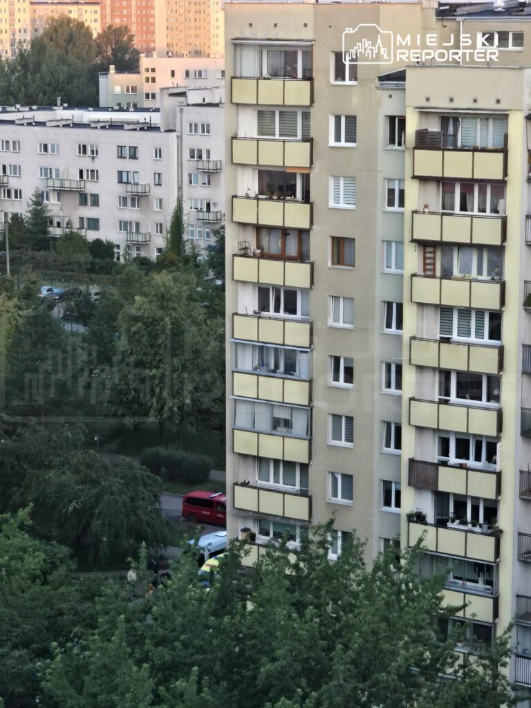 Blok mieszkalny z żółtymi balkonami otoczony drzewami i zaparkowanymi samochodami na ulicy.