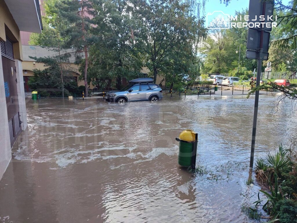 Ulica zalana wodą, na której unosi się samochód, a w tle widać drzewa i budynki mieszkalne.