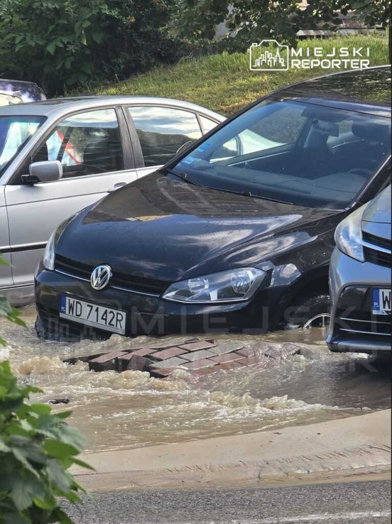 Czarny samochód osobowy unosi się na wodzie w zalanym miejscu, obok stoją inne pojazdy.