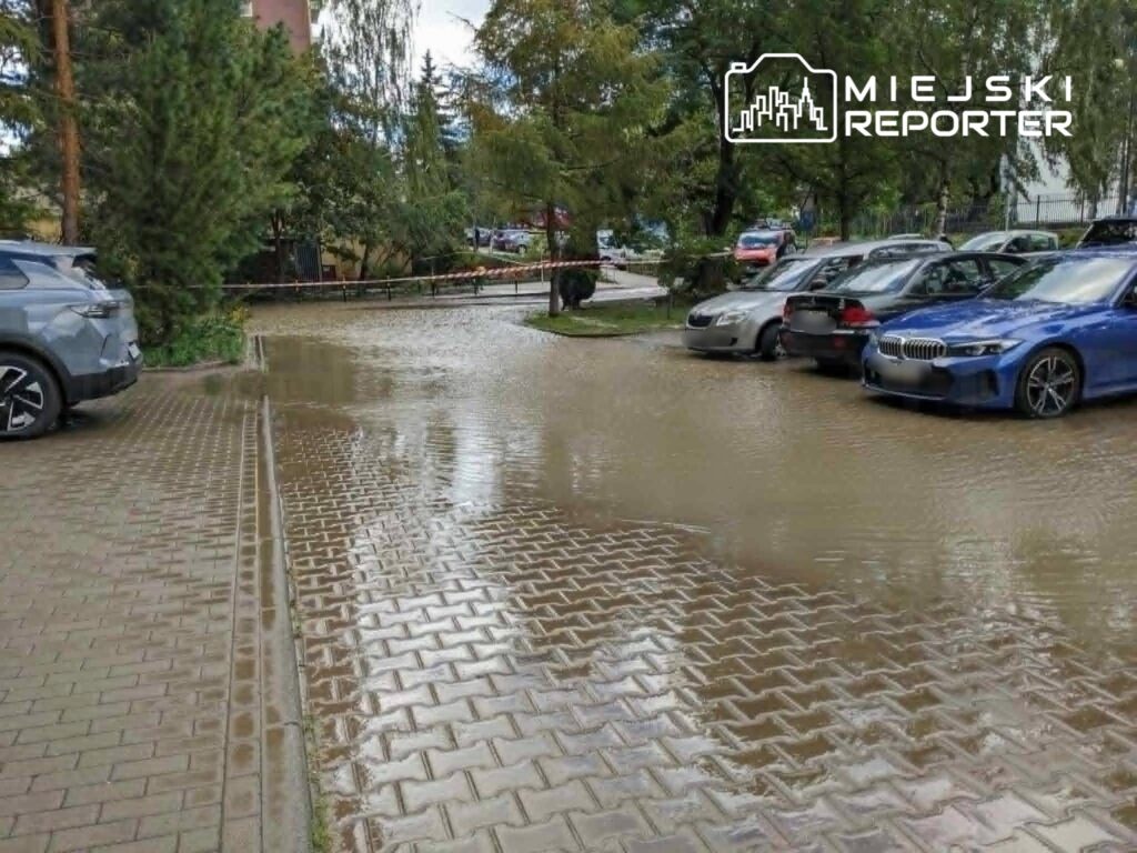 Zalany parking z samochodami, woda pokrywa kostkę brukową w otoczeniu drzew i budynków.