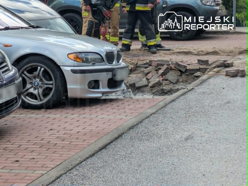 Czterech strażaków stojących obok uszkodzonej nawierzchni drogi, z której wydobywa się woda.