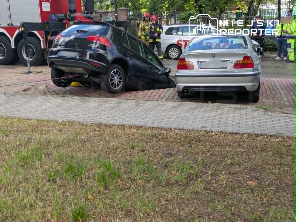 Czarny samochód osobowy unosi się na krawężniku, obok stoi srebrny sedan, a w tle widać strażaków.