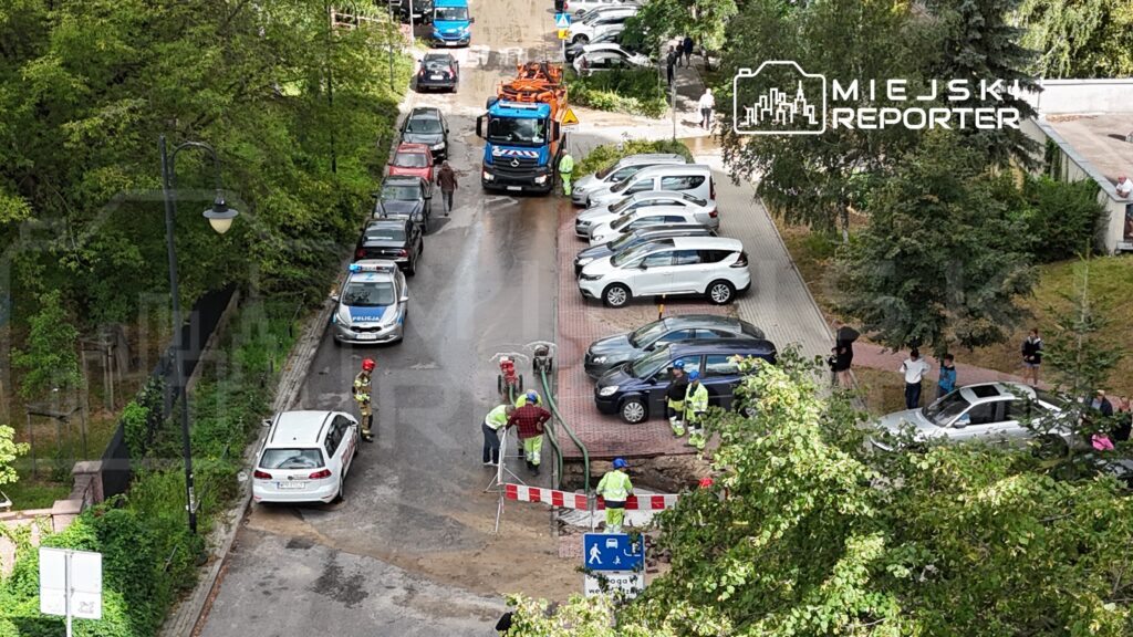 Grupa pracowników w odblaskowych kamizelkach naprawia nawierzchnię drogi, obok stoi policyjny radiowóz.