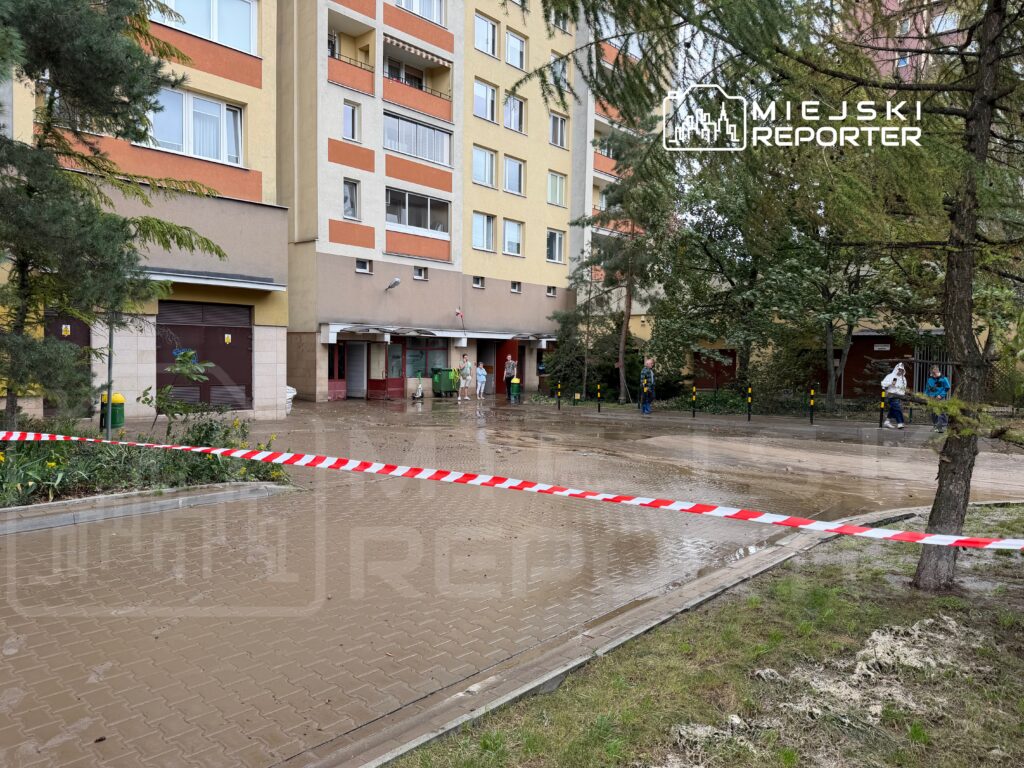 Na chodniku przed budynkiem mieszkalnym znajduje się błoto, a teren jest ogrodzony taśmą ostrzegawczą. W tle widać przechodniów oraz zieleń drzew.