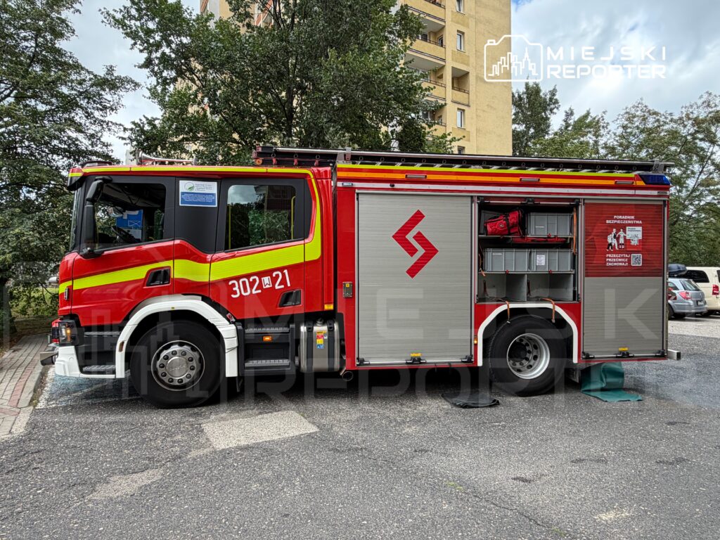 Czerwony wóz strażacki zaparkowany na osiedlowym parkingu, z otwartą zabudową tylną, w tle drzewa i budynki mieszkalne.