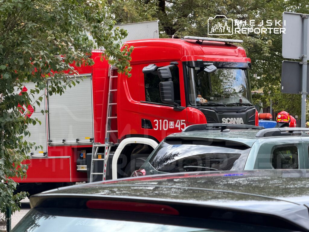 Czerwony wóz strażacki z drabiną stoi na ulicy, otoczony przez zaparkowane samochody.