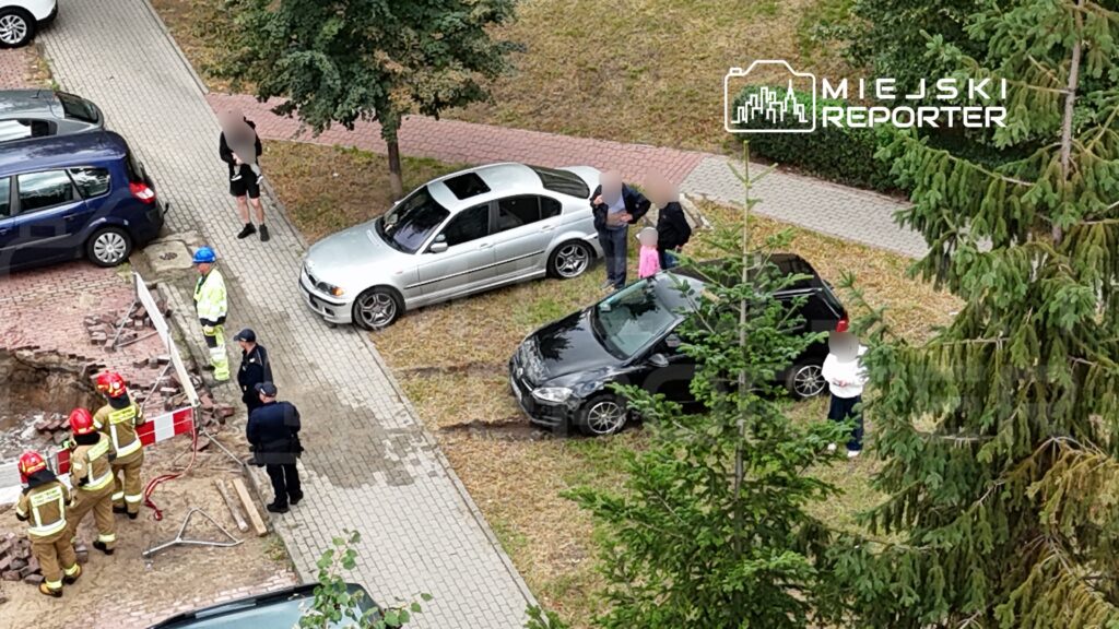 Grupa strażaków w mundurach i kaskach pracuje przy wykopie w chodniku, obok zaparkowane samochody.
