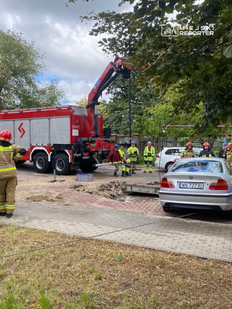 Grupa strażaków oraz pracowników w kamizelkach odblaskowych pracuje przy usuwaniu zniszczonej nawierzchni drogi.