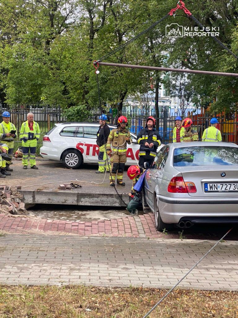 Grupa ratowników w kaskach i odblaskowych strojach pracuje przy usuwaniu zniszczonego fragmentu chodnika obok samochodu.