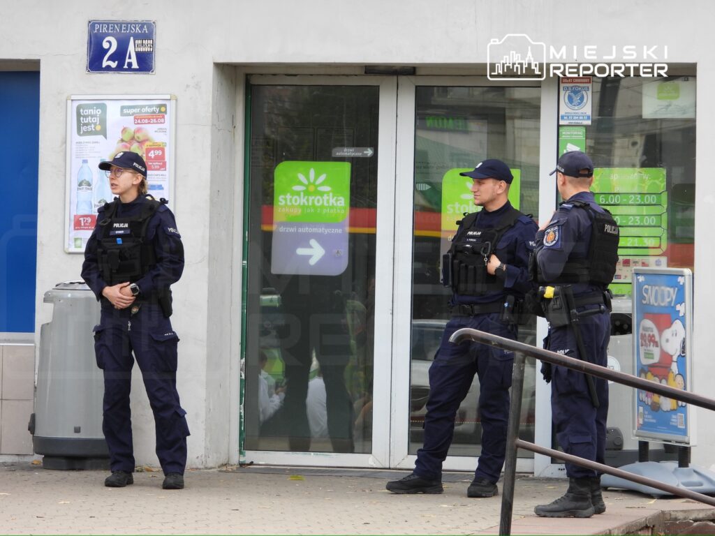 Trzech policjantów w mundurach stoi przed sklepem, jeden z nich rozmawia z kolegami, a w tle widoczne są reklamy.