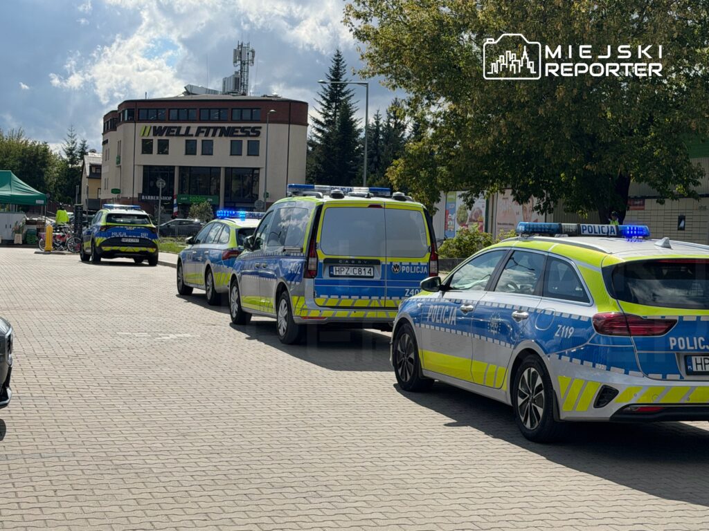 Policyjne samochody z włączonymi sygnałami świetlnymi zaparkowane przed budynkiem z napisem „WELLFITNESS”.