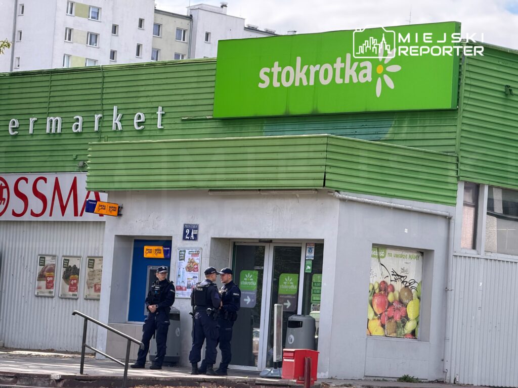 Czterech policjantów rozmawia przed supermarketem Stokrotka, w tle widoczne budynki mieszkalne.