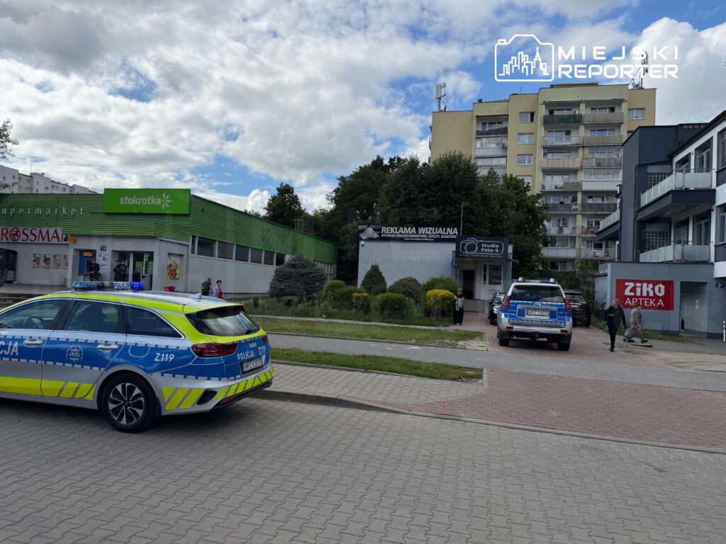 Policyjny radiowóz z włączonymi sygnałami świetlnymi stoi przed supermarketem, obok niego kilka osób na chodniku.