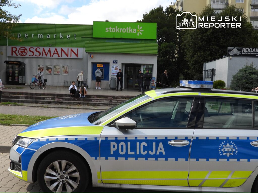 Policyjny radiowóz zaparkowany przed supermarketem, obok grupy osób czekających na chodniku.
