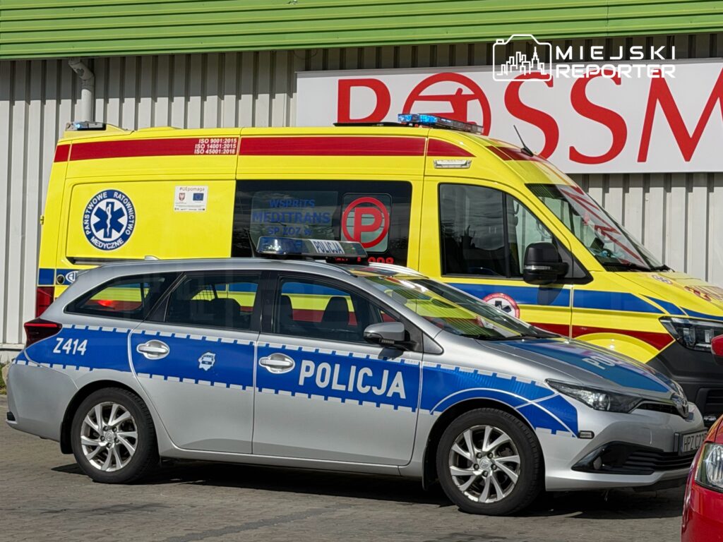 Policyjny samochód osobowy z niebieskimi oznaczeniami stoi obok karetki pogotowia w kolorze żółtym i czerwonym.