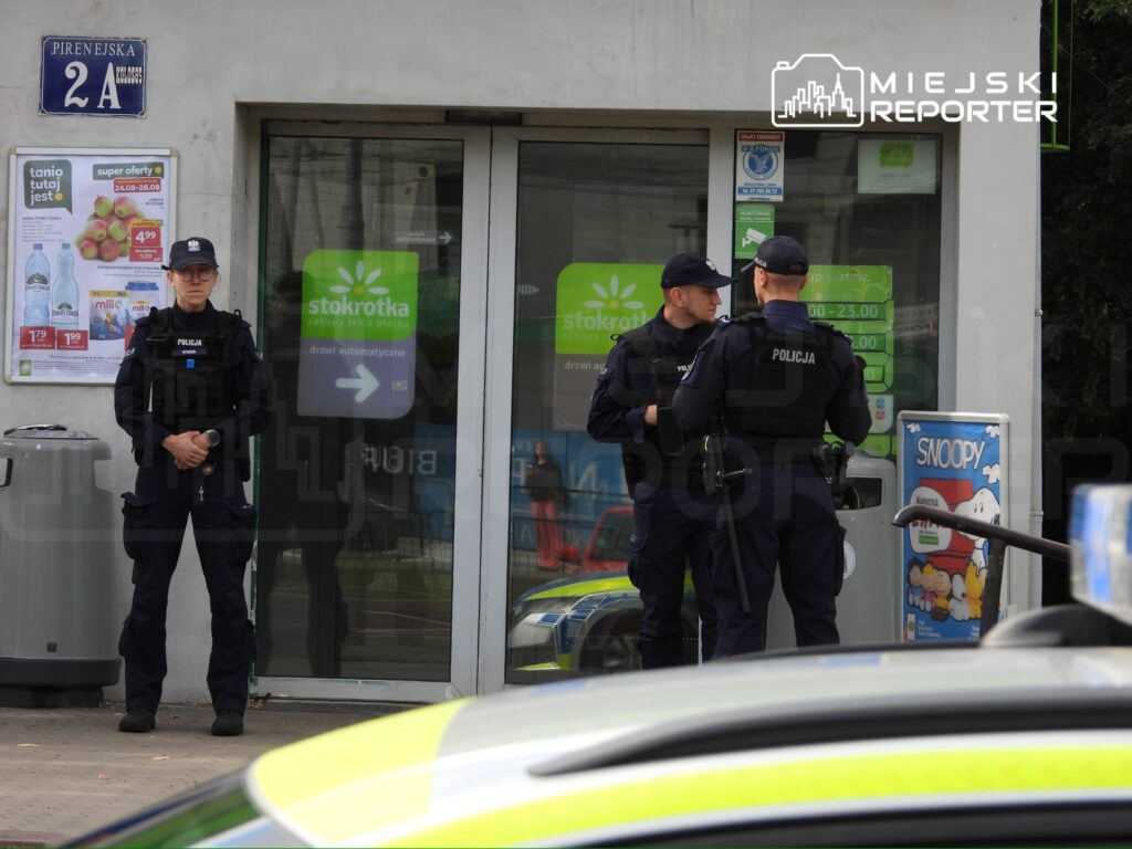Trzech policjantów w mundurach rozmawia przed sklepem, jeden z nich stoi na straży.