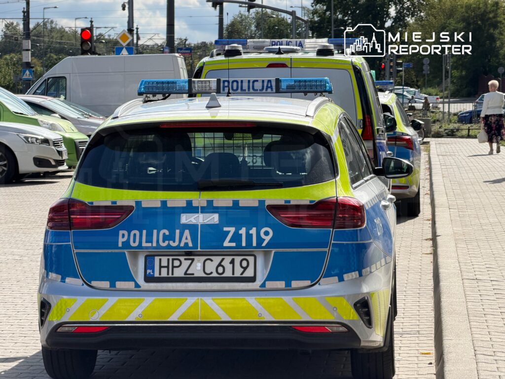 Radiowóz policyjny z niebieskimi i żółtymi znakami zatrzymany na ulicy, obok innych pojazdów w ruchu.