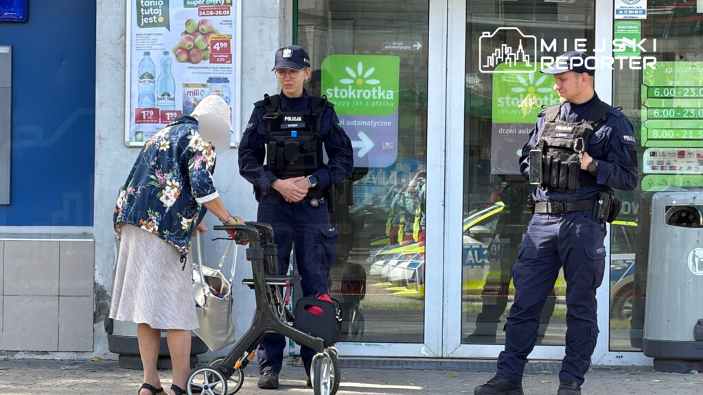 Starsza kobieta z wózkiem inwalidzkim rozmawia z dwoma policjantami przed sklepem spożywczym.