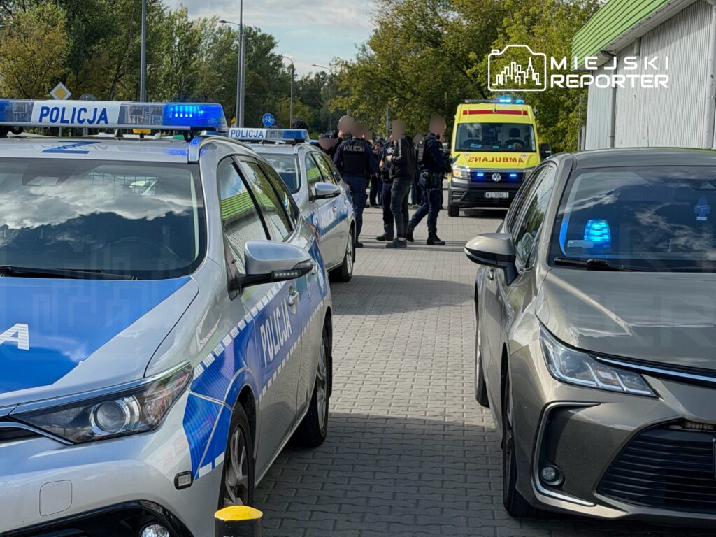 Policjanci w mundurach rozmawiają przy radiowozach na parkingu, w tle stoi karetka pogotowia z włączonym sygnałem.