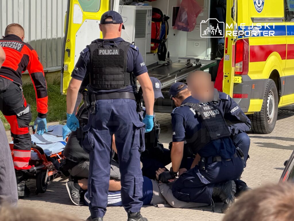 Grupa policjantów oraz ratowników medycznych udziela pomocy mężczyźnie leżącemu na ziemi obok karetki.